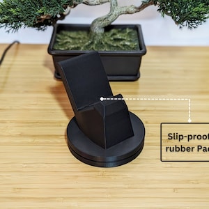May include: A black phone stand with a slip-proof rubber pad on the bottom. The stand is angled to hold a phone upright.