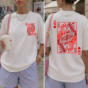 Op de afbeelding: Wit T-shirt met een rood Queen of Hearts speelkaartontwerp op de voor- en achterkant. De voorkant heeft een enkele kaart, terwijl de achterkant twee kussende koninginnen laat zien. Het shirt heeft korte mouwen en een losse pasvorm.