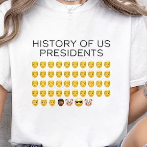 Pu&ograve; includere: Maglietta bianca con il testo "HISTORY OF US PRESIDENTS" e una griglia di emoji di facce gialle, tra cui un emoji di faccia nera, un emoji con occhiali da sole e un emoji di clown.