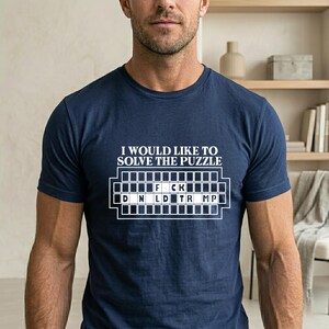 Pu&ograve; includere: T-shirt blu navy con la scritta "I WOULD LIKE TO SOLVE THE PUZZLE" sopra un gioco di parole che compone "F*CK D*N*LD TR*MP" in bianco. La maglietta &egrave; a maniche corte.