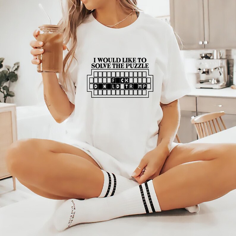 Pu&ograve; includere: T-shirt bianca con la scritta "I WOULD LIKE TO SOLVE THE PUZZLE" sopra una griglia di puzzle con le lettere "D N L D F C K TR MP". La persona indossa calzini bianchi con strisce nere e tiene in mano un bicchiere di caff&egrave; freddo.