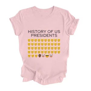 Pu&ograve; includere: Una maglietta rosa con il testo "HISTORY OF US PRESIDENTS" e una griglia di emoji gialli con facce sorridenti, tra cui un emoji nero con occhiali da sole, un emoji di clown e un emoji di clown con occhiali da sole.