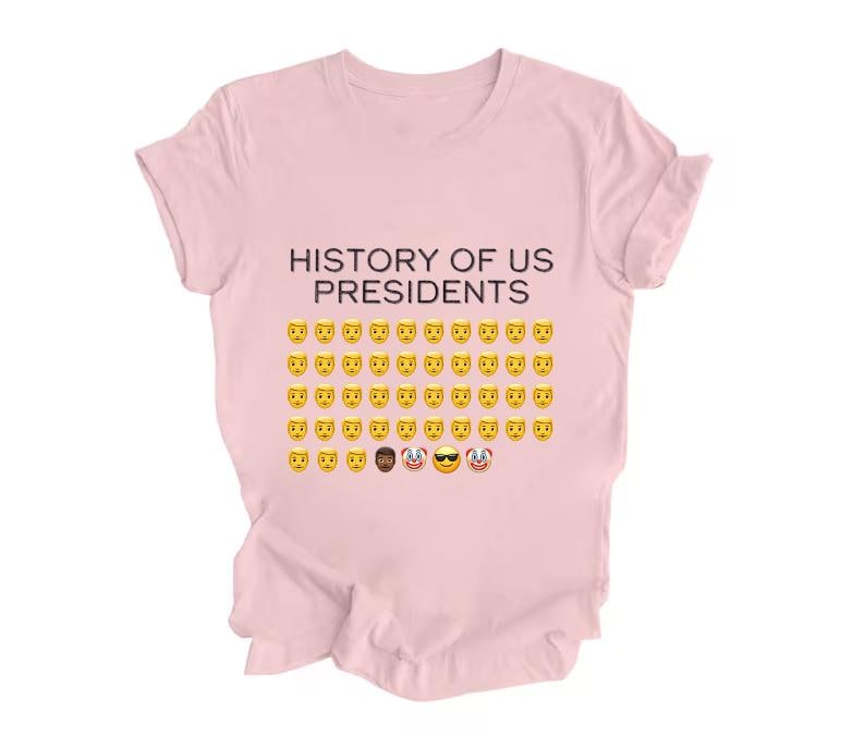 Pu&ograve; includere: Una maglietta rosa con il testo "HISTORY OF US PRESIDENTS" e una griglia di emoji gialli con facce sorridenti, tra cui un emoji nero con occhiali da sole, un emoji di clown e un emoji di clown con occhiali da sole.