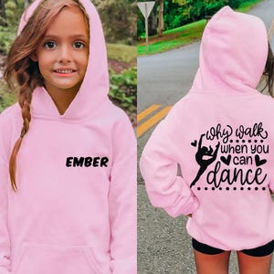 Puede incluir: Una sudadera con capucha rosa claro con el nombre "EMBER" impreso en negro en la parte delantera. La parte trasera de la sudadera con capucha presenta un gráfico de bailarina y el texto "why walk when you can dance" en negro.