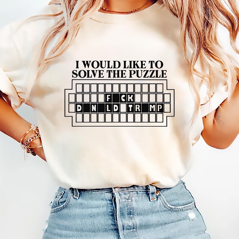 Pu&ograve; includere: Felpa color crema con la scritta "I WOULD LIKE TO SOLVE THE PUZZLE" sopra una griglia di puzzle. La griglia dice "F CK" e "D N L D TR MP". La felpa &egrave; abbinata a pantaloncini di jeans blu.