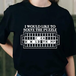 Pu&ograve; includere: T-shirt nera con la scritta "I WOULD LIKE TO SOLVE THE PUZZLE" sopra una griglia di puzzle. La griglia contiene le lettere "F CK" e "D N L D TR MP". Un capo d'abbigliamento casual.
