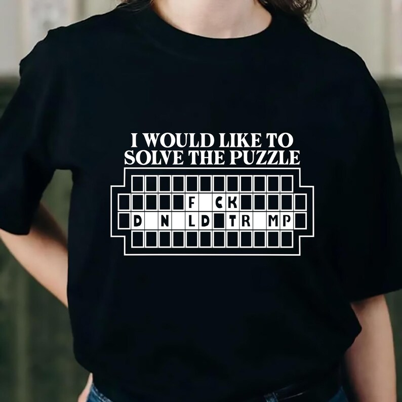 Pu&ograve; includere: T-shirt nera con la scritta "I WOULD LIKE TO SOLVE THE PUZZLE" sopra una griglia di puzzle. La griglia contiene le lettere "F CK" e "D N L D TR MP". Un capo d'abbigliamento casual.