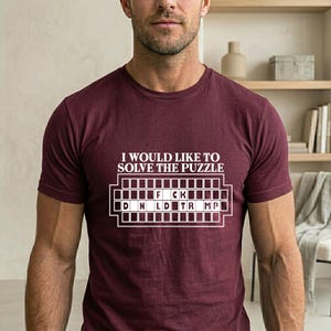 Pu&ograve; includere: T-shirt bordeaux con la scritta "I WOULD LIKE TO SOLVE THE PUZZLE" sopra un motivo a cruciverba con le lettere "F CK" e "D N L D TR MP". Girocollo.