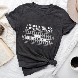 Pu&ograve; includere: T-shirt grigio scuro con la scritta "I would like to solve the puzzle" e un gioco di parole che scrive "F*ck D N L D TR MP" in bianco. La maglietta &egrave; annodata in basso. Un cappello, una tazza di caff&egrave; e un libro sono anche nell'immagine.