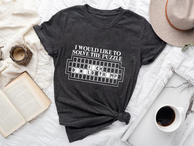 Pu&ograve; includere: T-shirt grigio scuro con la scritta "I would like to solve the puzzle" e un gioco di parole che scrive "F*ck D N L D TR MP" in bianco. La maglietta &egrave; annodata in basso. Un cappello, una tazza di caff&egrave; e un libro sono anche nell'immagine.