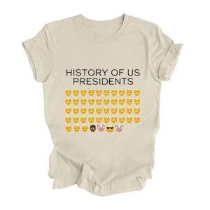 Pu&ograve; includere: Una maglietta beige con la scritta "HISTORY OF US PRESIDENTS" stampata sul davanti. Il testo &egrave; seguito da una griglia di faccine emoji gialle.
