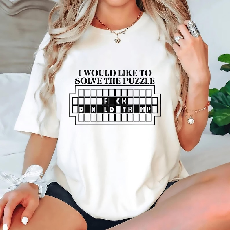 Pu&ograve; includere: T-shirt bianco sporco con la scritta nera "I WOULD LIKE TO SOLVE THE PUZZLE". Sotto, una grafica di un gioco di parole con le lettere F, C, K, D, N, L, D, T, R e MP.