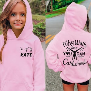 Op de afbeelding: Een lichtroze hoodie met een voorzak en capuchon. De voorkant heeft een zwarte afbeelding van gymnasten en de naam "KATE". De achterkant van de hoodie heeft de tekst "Why Walk When You Can Cartwheel" in het zwart.