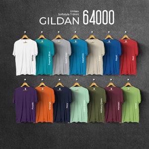 Pu&ograve; includere: Esposizione di magliette unisex Gildan 64000 Softstyle in vari colori, tra cui bianco, rosso e blu navy. Ogni maglietta &egrave; appesa a una gruccia di legno su uno sfondo grigio scuro. Il testo "GILDAN 64000" &egrave; visualizzato in alto.