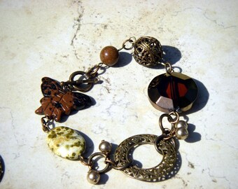 May include: A brown and black butterfly charm, a brown and white speckled stone, a clear red and brown faceted glass bead, a gold metal bead with an openwork design, and a gold metal clasp make up this bracelet.