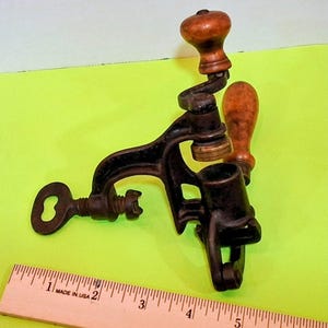 May include: An antique, cast iron meat grinder with a wooden handle and a key-shaped clamp. The grinder is black with a brass fitting and a wooden handle. A wooden ruler with "MADE IN USA" is in the foreground.