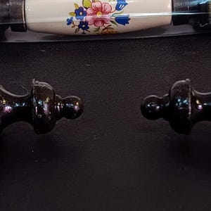 May include: Two white ceramic cabinet knobs with black metal accents. The knobs have a simple, oval shape and are attached to a decorative metal base.