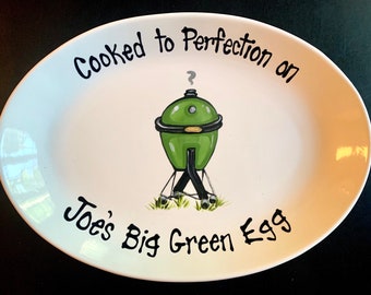 May include: White oval ceramic serving platter with black lettering that reads "Cooked to Perfection on Joe's Big Green Egg". A green and black grill with smoke coming out of the top is in the center of the platter.