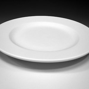 May include: A white ceramic dinner plate with a slightly raised rim.