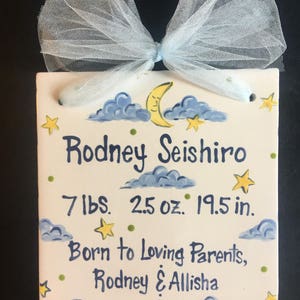 May include: A white ceramic tile with a blue and yellow painted design of clouds, stars, and a crescent moon. The tile has the text "Rodney Seishiro 7 lbs. 2.5 oz. 19.5 in. Born to Loving Parents, Rodney & Allisha" written in black.