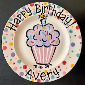 May include: A round ceramic plate with a cream-colored base, featuring a hand-painted purple cupcake with a single candle in the center. The rim is decorated with colorful polka dots. The text reads Happy Birthday! July 25th Avery in black.