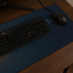 Könnte beinhalten: Eine schwarze Tastatur, Maus und Mauspad auf einem Holztisch. Das Mauspad ist dunkelblau mit einem Leiterplattendesign und dem Aufdruck "Eng". Die Tastatur ist eine Standardgröße mit schwarzen Tasten.