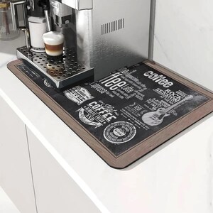 Puede incluir: Una máquina de espresso de acero inoxidable con una taza de café en la bandeja de goteo. Una alfombrilla negra y marrón con temática de café con texto y gráficos se encuentra en la encimera. La alfombrilla tiene las palabras "coffee", "espresso" y "latte".