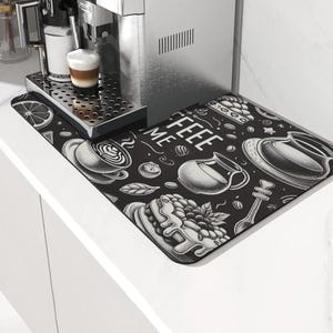 Könnte beinhalten: Eine Espressomaschine aus Edelstahl mit einer Tasse Kaffee auf der Tropfschale. Eine schwarze Matte mit Kaffeemotiven und den Worten "Coffee Me" befindet sich im Vordergrund. Die Matte zeigt Kaffeetassen, Gebäck und Kaffeebohnen.