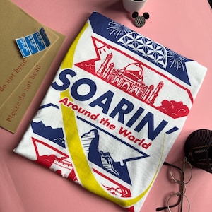 May include: White t-shirt with a blue and red graphic design that says "SOARIN' Around the World". The graphic features a stylised image of a building with minarets and a yellow crescent moon.