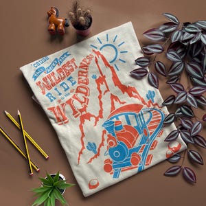 May include: A beige t-shirt featuring a graphic of a train travelling through a mountain range. The text on the shirt reads "'cause this here's the WILDEST RIDE in the WILDERNESS".