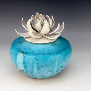 May include: A round ceramic jar with a vibrant turquoise blue glaze featuring a textured, mottled finish. The lid is topped with a sculpted, off-white ceramic flower with layered, pointed petals that resemble a succulent or lotus blossom.