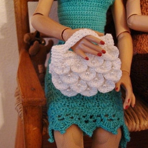 May include: A white crocheted handbag with a scalloped design. The bag is being held by a doll wearing a turquoise crocheted dress.