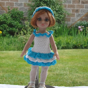 May include: A doll wearing a handmade crochet dress and headband. The dress is white with blue trim and ruffles. The doll has red hair and is wearing white tights and shoes. The background is a brick wall and green grass.