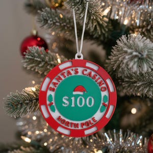 May include: A Christmas tree ornament shaped like a casino chip. The ornament is red, white, and green, with the text "SANTA'S CASINO NORTH POLE" and "$100". A Santa hat is in the center.