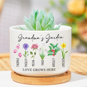 Può includere: Vaso in ceramica bianca con il testo "Grandma's Garden" e una pianta grassa. Il vaso è decorato con un motivo floreale con i nomi Thomas, Paisley, Micah, Paige, Kinsley e Lincoln scritti sotto ogni fiore.