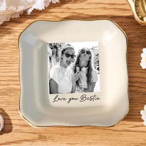 May include: A cream-colored ceramic trinket dish with a gold trim, featuring a black and white photo of two women. The phrase "Love you Bestie" is written below the photo. The dish is on a wooden surface.