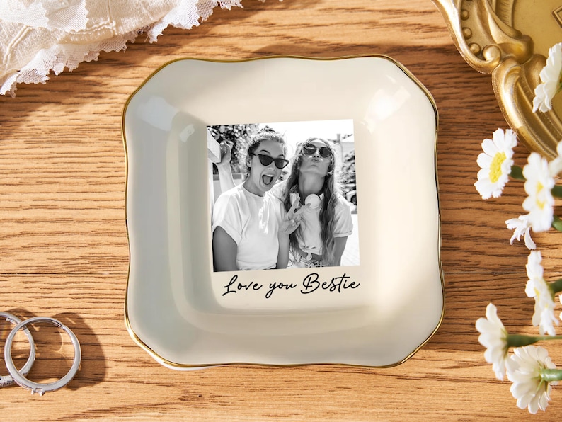 May include: A cream-colored ceramic trinket dish with a gold trim, featuring a black and white photo of two women. The phrase "Love you Bestie" is written below the photo. The dish is on a wooden surface.