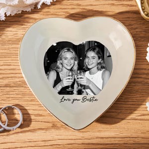 May include: Heart-shaped trinket dish with a black and white photo of two women toasting. The dish is cream-colored with a gold trim and the words "Love you Bestie" are written on the front. Two rings are next to the dish.