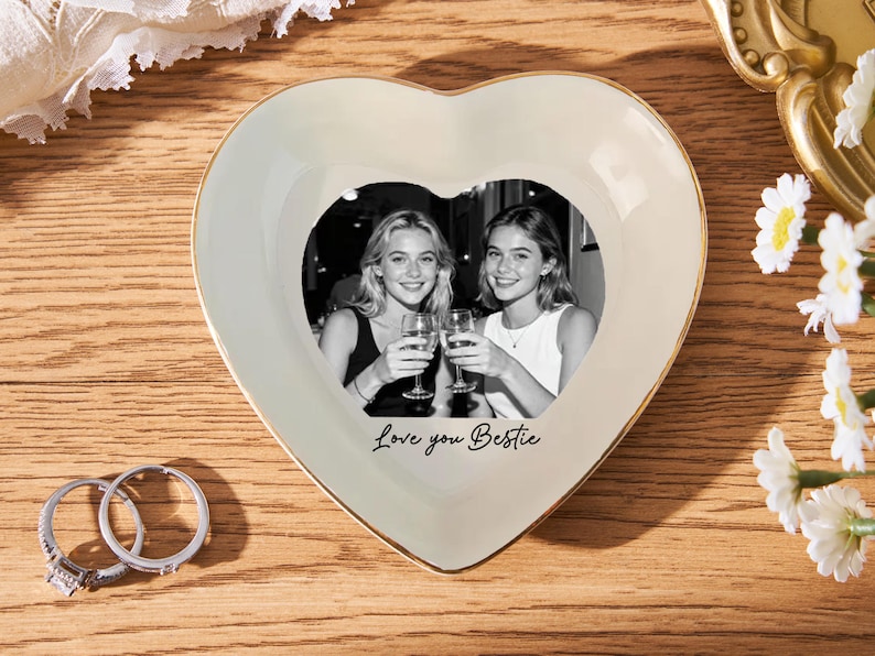 May include: Heart-shaped trinket dish with a black and white photo of two women toasting. The dish is cream-colored with a gold trim and the words "Love you Bestie" are written on the front. Two rings are next to the dish.