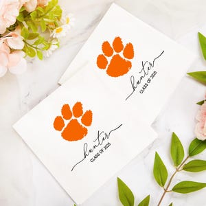 May include: Two white cards with an orange paw print and the name "Hunter" in cursive, with "CLASS OF 2025" below. The cards are surrounded by flowers and greenery.