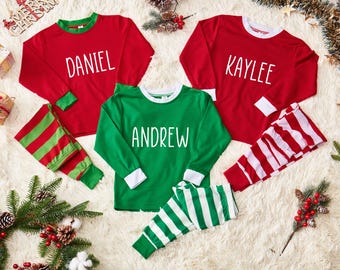 May include: Red and green personalized pajama sets with names printed on the shirts. The long-sleeved shirts have matching striped pajama pants. The names "DANIEL", "KAYLEE", and "ANDREW" are displayed on the shirts. Festive holiday theme.