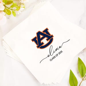 May include: White paper napkins with a navy blue and orange university logo and the name "Olivia" in cursive, along with "CLASS OF 2025". The napkins are stacked and surrounded by flowers and greenery.