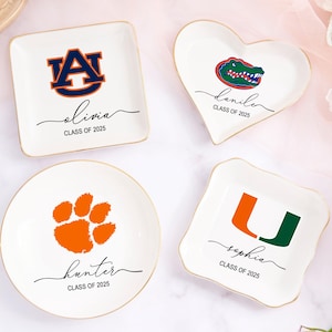 May include: Four white ceramic trinket dishes with gold rims. Each dish features a different college logo and a name with "CLASS OF 2025". The shapes include a square, heart, round, and a square with wavy edges. The logos are in orange, green, and navy blue.