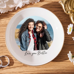 May include: A white ceramic plate with a gold rim features a photo of two smiling people. The plate has the text "Love you Bestie" in black script. Two rings and flowers are also visible.