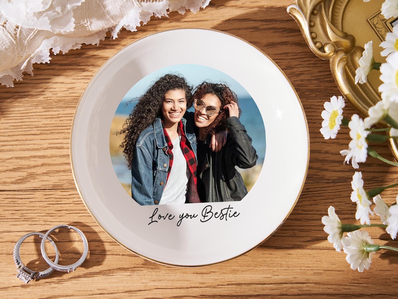 May include: A white ceramic plate with a gold rim features a photo of two smiling people. The plate has the text "Love you Bestie" in black script. Two rings and flowers are also visible.