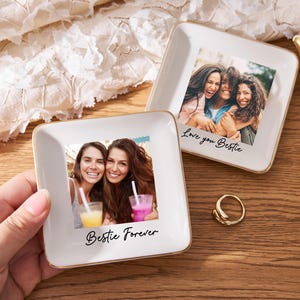 May include: Two square, white ceramic trinket dishes with gold trim, each featuring a photograph of smiling women. One dish has "Bestie Forever" and the other "Love you Bestie" in script. A gold ring and flowers are also present.