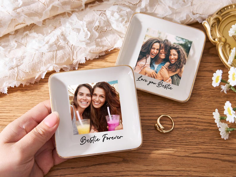 May include: Two square, white ceramic trinket dishes with gold trim, each featuring a photograph of smiling women. One dish has "Bestie Forever" and the other "Love you Bestie" in script. A gold ring and flowers are also present.