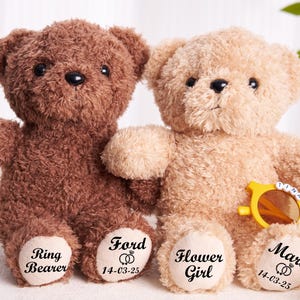 Puede incluir: Dos osos de peluche, uno marrón y otro beige, con mensajes personalizados en sus patas. El oso marrón tiene "Ring Bearer 14-03-25" en su pata, y el oso beige tiene "Flower Girl 14-03-25" en su pata. El oso beige lleva gafas de sol.