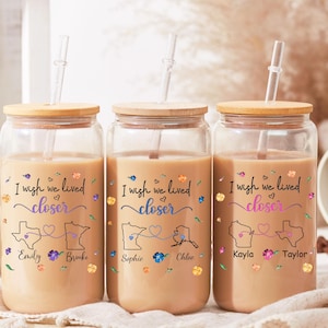 May include: Three glass tumblers with wooden lids and straws, each filled with a light brown liquid. The tumblers are decorated with the phrase "I wish we lived closer", state outlines, and names. Floral accents in various colors surround the text.