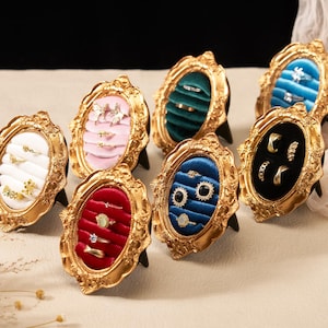 May include: A collection of gold-coloured, ornate framed ring displays. Each frame has a velvet interior in colours such as white, pink, green, red, blue, and black. The frames, each holding rings, are set on small black stands, showcasing jewellery.
